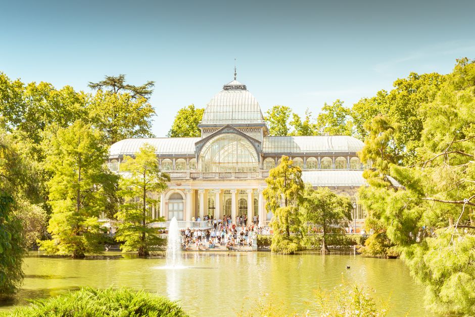 A large, ornate glass conservatory situated in Crystal Palace Park surrounded by lush green trees and a pond with a central fountain. The conservatory features a domed roof with intricate metalwork and wide windows, and it appears well-maintained with clean glass panels. The scene is brightly lit with natural sunlight, highlighting the vibrant greenery and the building's elegant architecture. Visitors are visible on the steps leading to the entrance, emphasizing the park's popularity for leisurely visits and sightseeing. The overall atmosphere reflects a serene, well-kept outdoor space ideal for family outings and outdoor relaxation, complementing the guide for surface cleaning and park maintenance provided by Carpet Cleaners SE19.
