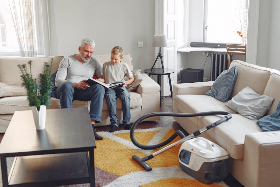 A living room with light-colored walls and large windows allowing natural light to fill the space. A grey sofa is positioned along the wall, with a young boy and an older man sitting on it, both engaged with a book. The room features a wooden floor covered partially by a patterned area rug in shades of yellow, orange, and beige. Nearby, a vacuum cleaner with a grey and white body and a black hose rests on the floor, indicating recent surface cleaning. To the left, a black coffee table holds a white vase with green leafy plants. Adjacent to the sofa, a small round side table with a modern lamp and a radiator under the window add to the cozy atmosphere. The room displays a tidy appearance with clean cushions and minimal clutter, suggesting routine domestic cleaning and surface sanitisation, consistent with the focus of the Crystal Palace Park carpet cleaning guide for families by Carpet Cleaners SE19.