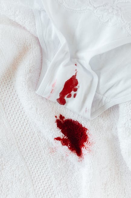 A person's hand holding a white fabric cloth stained with bright red blood splatters, with a light pink background. The cloth appears to be part of a garment or cleaning cloth, and the image emphasize
