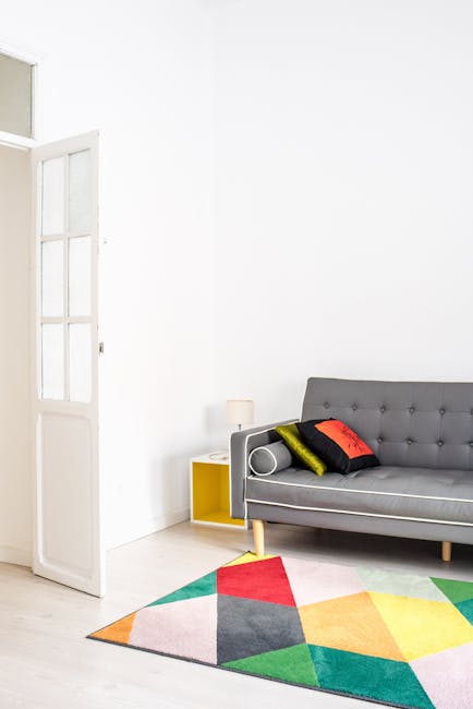 A bright, minimalist living room featuring a light-colored wooden floor partially covered by a multicolored geometric rug with shades of red, yellow, green, and gray. Against a plain white wall, there